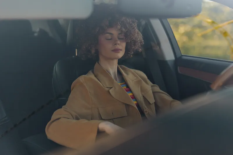 A woman driver rests with eyes closed in a warm retro car interior, suggesting calm travel, self care, and a quiet moment during a casual road journey at golden hour.