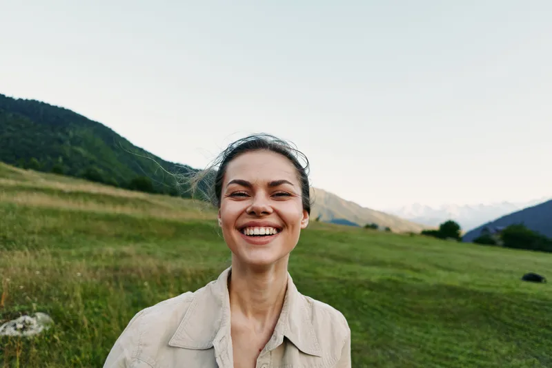 Bright smiling young woman stands in a green countryside field, wearing a light beige jacket, enjoying warm sun and open space, a candid moment of everyday wellbeing