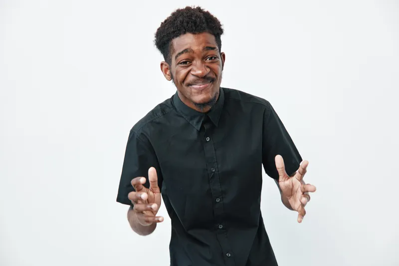 Young black man with dark skin wearing black shirt smiling and gesturing with hands on white background. African ethnicity, casual style, positive expression and confident pose.