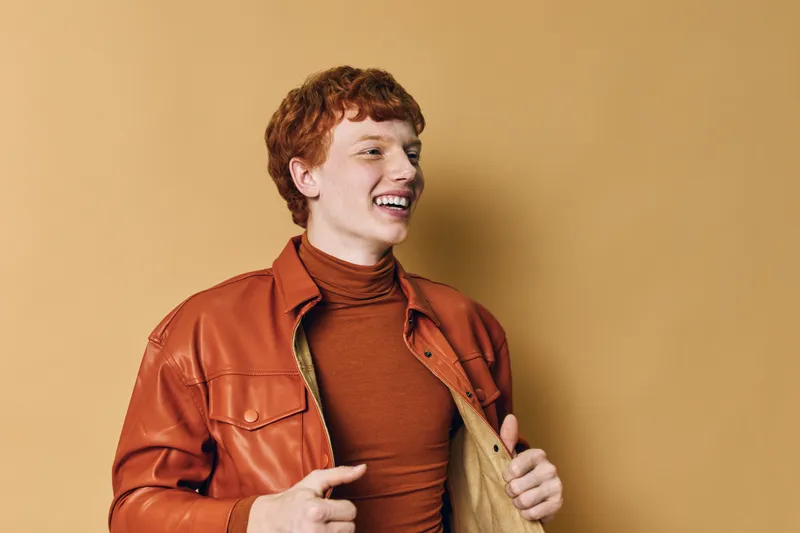 Young man with curly hair wearing brown leather jacket and turtleneck stands smiling on beige background. Casual style, confident expression, fashion portrait with warm tones.