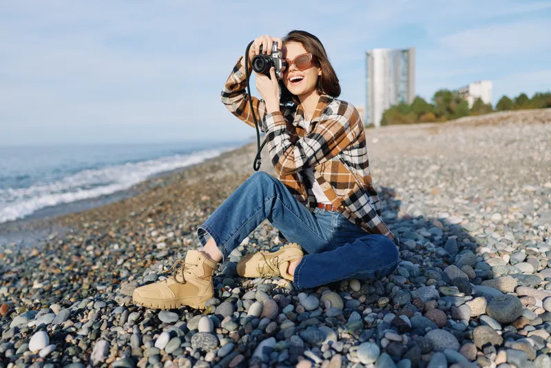 Outdoor scene of a woman on a pebble beach with a camera, wearing plaid shirt and jeans, enjoying daylight by the sea, capturing candid moments and natural scenery with a focused lens.