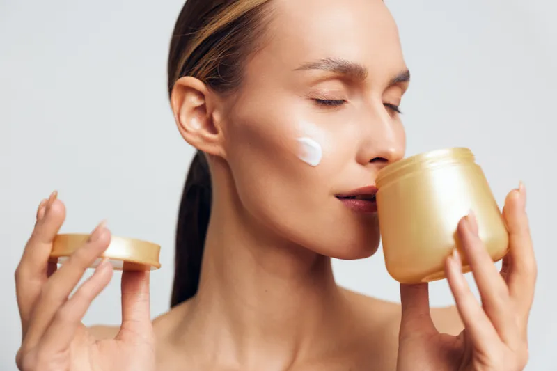 A young woman applies cream on her cheek while holding an open jar of skincare product close to her face with eyes closed in a bright studio. Concept for beauty and self-care.