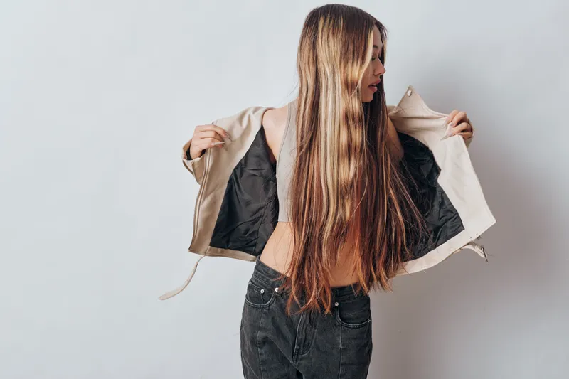 Young woman with long hair wearing black jeans and beige jacket open in studio against light background. Fashion, beauty, casual style and portrait for clothing and lifestyle.