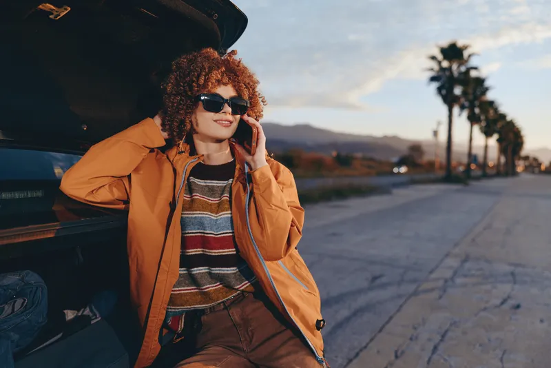 Woman lifestyle portrait by car at sunset, street background, wearing sunglasses, warm coat and striped sweater, holding phone to ear, curly hair in wind, casual fashion travel moment.