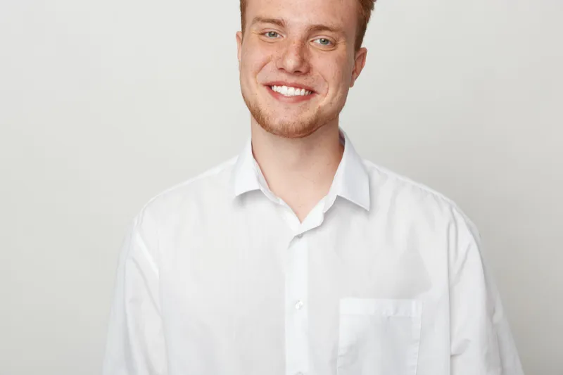 Smiling young man with red hair and beard wearing white shirt stands against plain light background. Portrait of happy male model with casual style and positive facial expression.