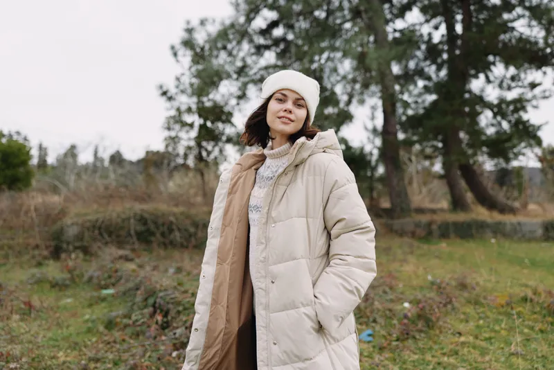 Woman in a beige puffer coat, white beanie and scarf stands in a chilly field, natural light and relaxed pose, autumnal landscape behind, casual style and cozy outdoor look.