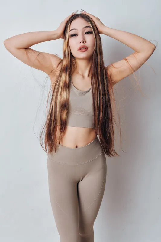 Young woman posing with hands on head in beige sportswear against white background, showing slim figure and long hair for fitness and lifestyle concept.