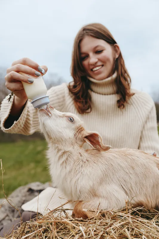 A young woman in a beige turtleneck sweater happily feeds a baby goat with a bottle outdoors on a cloudy day, capturing a warm lifestyle moment with animals.