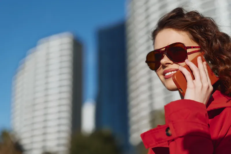 Stylish woman in a red coat talks on a smartphone in a bright urban setting, with glass skyscrapers rising behind her and a clear blue sky.
