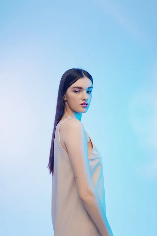 Young woman with long dark hair wears beige dress posing against blue background. Soft lighting highlights her beauty and fashion style. Concept for beauty and fashion.