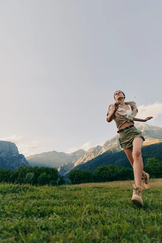 A young woman in hiking shorts and a tank top leaps across a grassy alpine meadow with distant mountains, expressing freedom and energy.