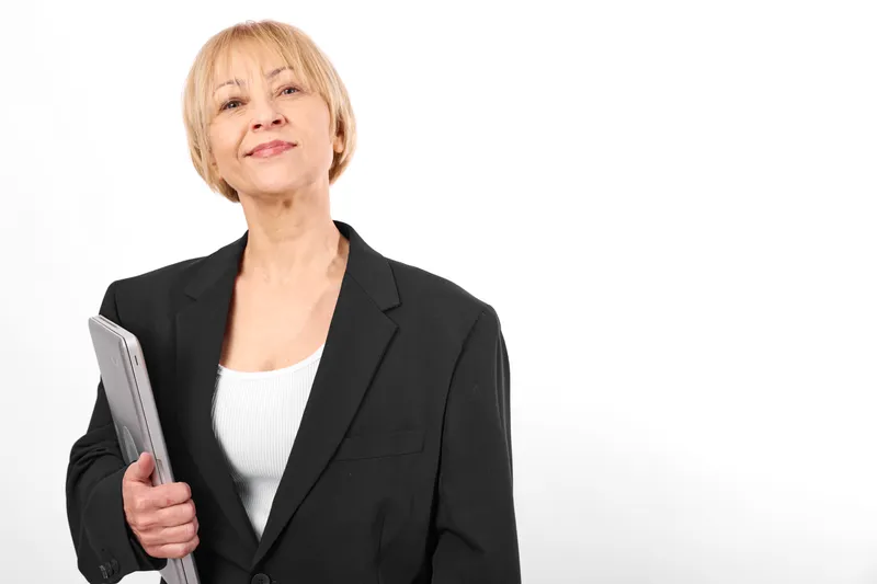 Senior woman business portrait in a black blazer, holding folder close to her body, calm smile, professional office look, isolated on white background, soft studio lighting.