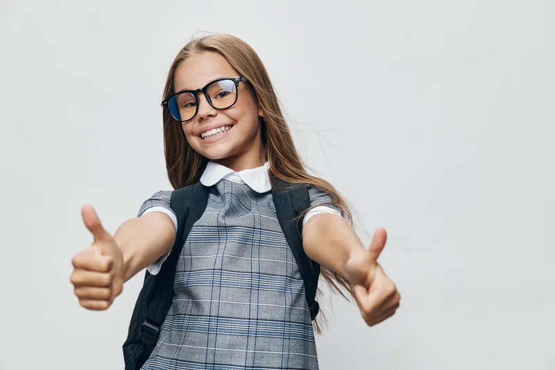 Girl child smiling with glasses giving thumbs up gesture wearing school uniform and backpack on gray background for education, success and positivity concept.