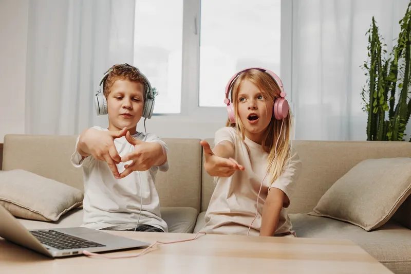 Two children wearing headphones enjoy music and dance together in a cozy living room with a laptop on the table. Concept of childhood fun and technology use.
