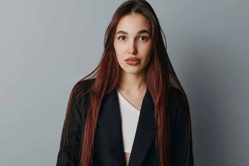 Woman with long straight hair and natural makeup wearing black jacket and white top looking at camera with neutral expression on gray background in studio portrait.