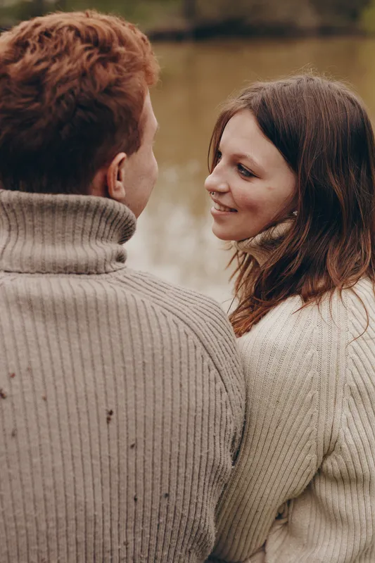 Warm candid moment of a couple outdoors, smiling and sharing a quiet conversation, cozy sweaters, gentle sunlight, natural tones, peaceful water backdrop, seasonal serenity, in a relaxed pose,