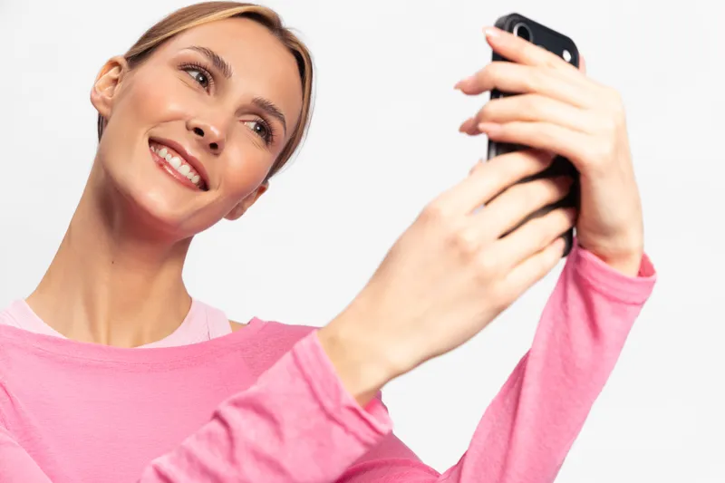 Smiling woman in pink long sleeve shirt takes selfie with smartphone on white background. Concept of happiness, technology, social media, communication, and casual lifestyle.