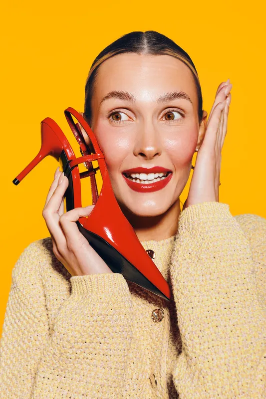 Young woman with shiny skin holds red high heel shoe near face, smiling with bright lipstick on yellow background. Concept for fashion, beauty, and stylish accessories.