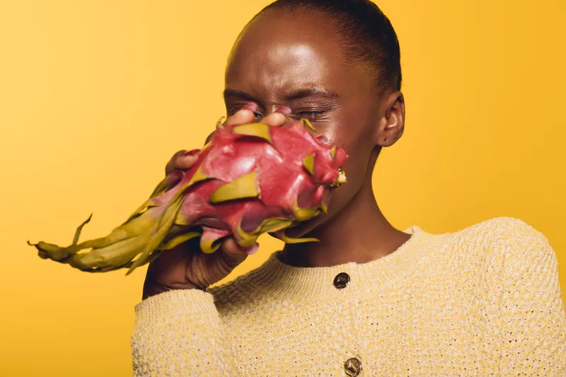 Young black woman with dark skin holds pink dragon fruit close to her face against yellow background. Concept of exotic fruit, healthy lifestyle, and vibrant natural beauty.