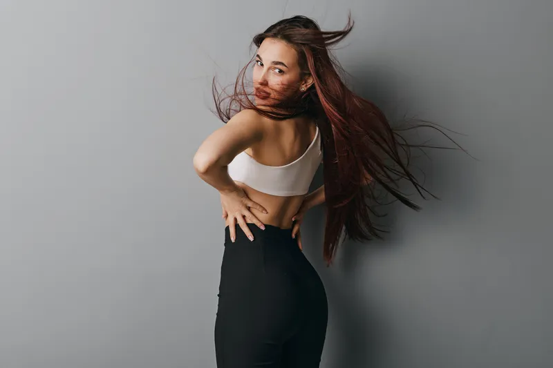 Young woman posing with long hair flowing wearing white top and black pants against gray background. Fashion, beauty, confident model with casual style and natural makeup.