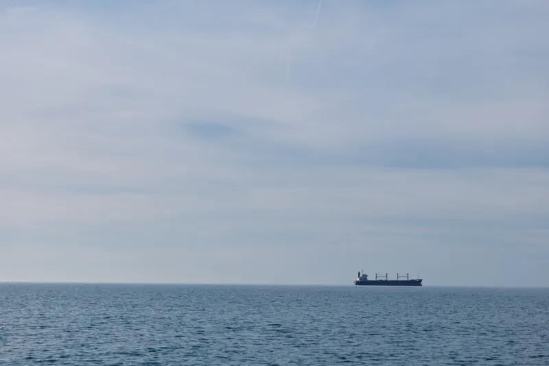 Calm sea with a distant cargo ship on the horizon under a soft cloudy sky, expansive water surface and a sense of travel and solitude across open ocean