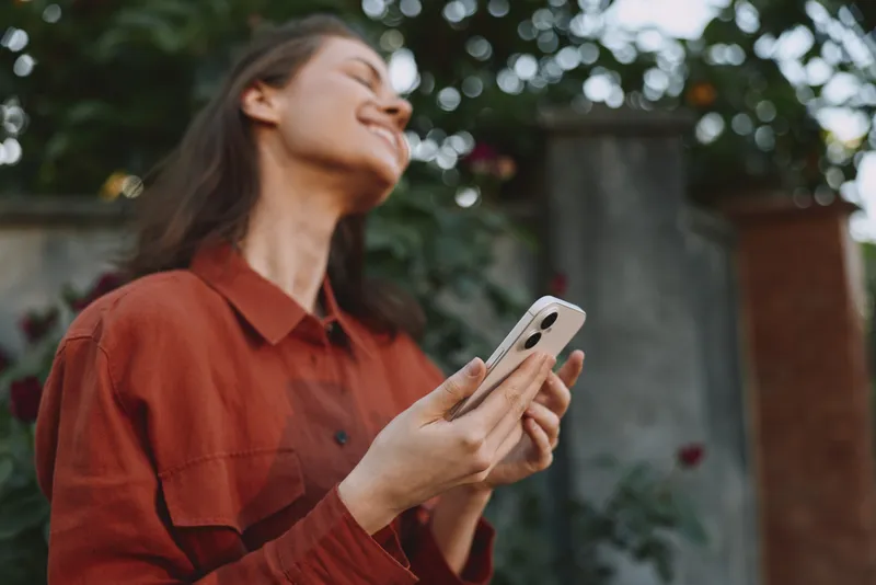 Smiling woman outdoors wearing a casual jacket holding a phone during daytime sunlight, capturing a candid lifestyle moment in a natural garden setting