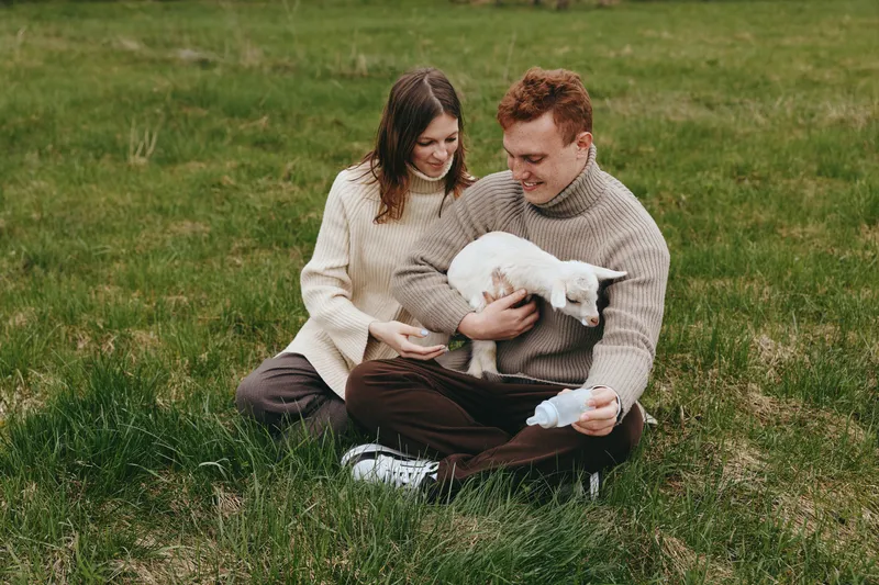 A young couple sits on green grass enjoying a playful moment with a white baby goat in a casual outdoor lifestyle scene during mild weather.