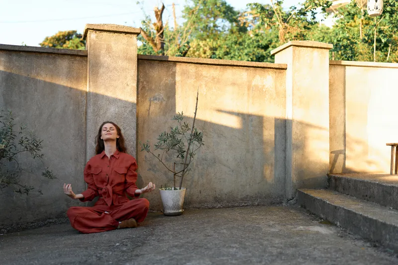 Woman meditating outdoors on a rooftop, sunlight highlights a red outfit, calm posture in a warm breeze against a simple urban backdrop
