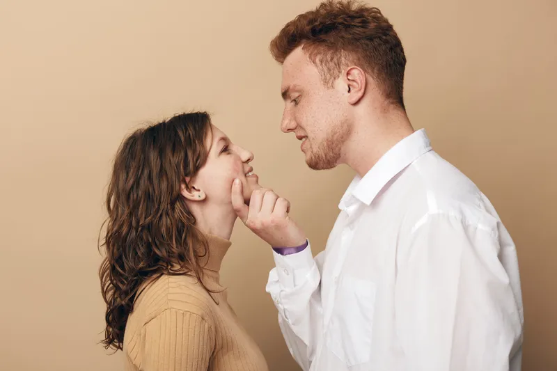 Young couple in casual clothes share intimate moment, man gently touches woman’s chin. Beige background creates warm atmosphere. Concept of love, romance, connection, and affection between partners.