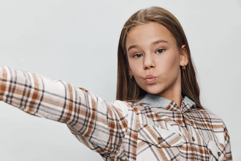 Teenage girl with long hair wearing plaid shirt making a selfie with pout lips on white background. Casual style, youth and social media concept.