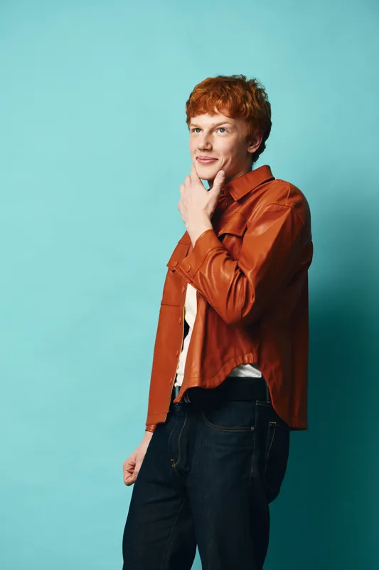 Young man with red hair wearing brown leather jacket and jeans standing against blue background thoughtful and looking away with hand on chin in casual pose.