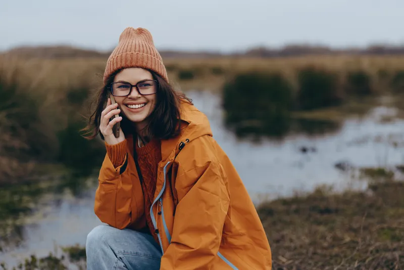 Smiling woman in orange jacket and knit hat talking on phone outdoors, lifestyle in nature landscape, cold winter air, soft daylight, relaxed candid mood