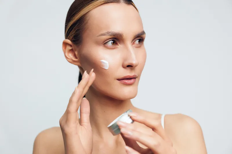 Young woman applying moisturizing cream on her face with fingers, clear skin, natural look, skincare routine, neutral background, focused expression, beauty care concept