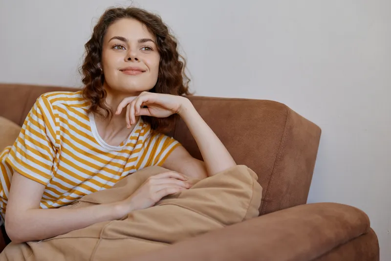 Cheerful woman relaxing on a sofa at home, wearing a yellow striped tee, resting under a soft blanket, warm natural light, candid lifestyle moment, cozy welcoming atmosphere