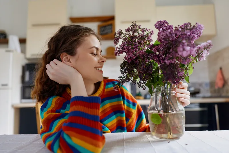 Young woman in a colorful striped sweater relaxes at home, smiling softly as she gazes at a vase of purple flowers on a bright table in a cozy kitchen