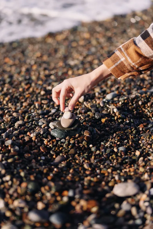 Person placing a pebble on a small rock stack along a pebbled beach with gentle waves in the background, a calm outdoor scene emphasizing balance and nature.