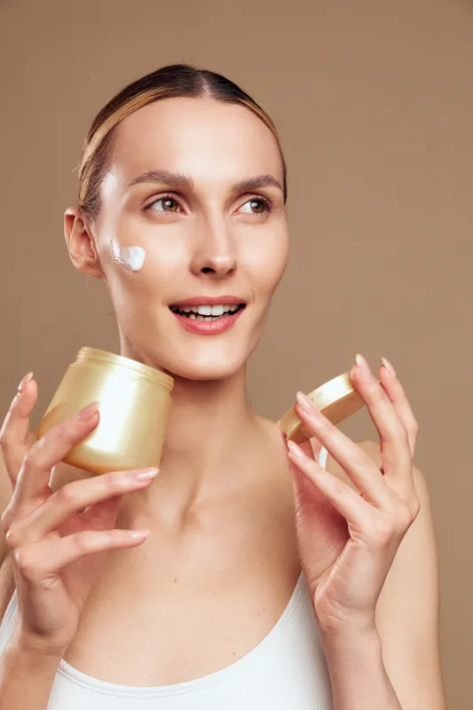 Smiling woman holding face cream jar and applying white cream on cheeks, close up against beige background, skincare routine and self care concept, studio lighting