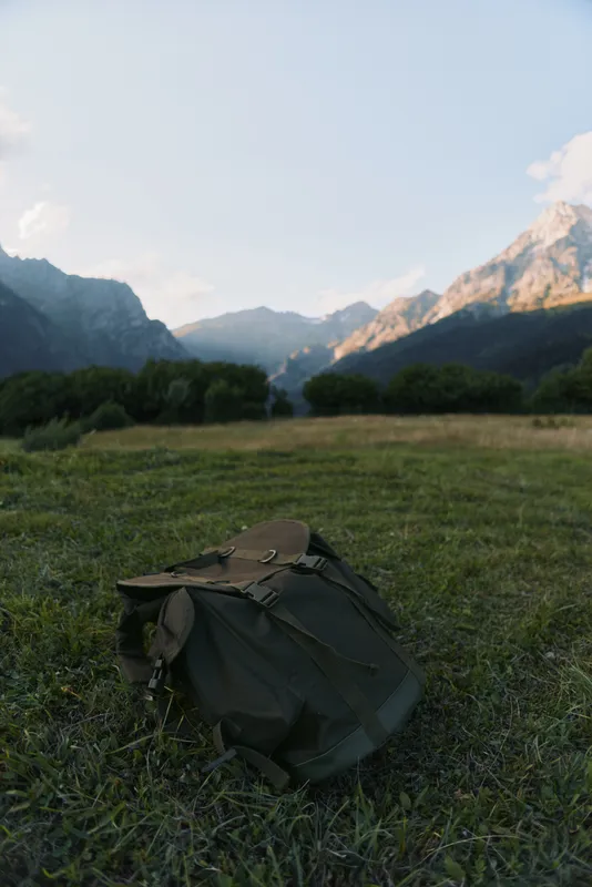 Green backpack lies on grass field in mountain valley during late afternoon with clear sky and distant peaks illuminated by warm sunlight.