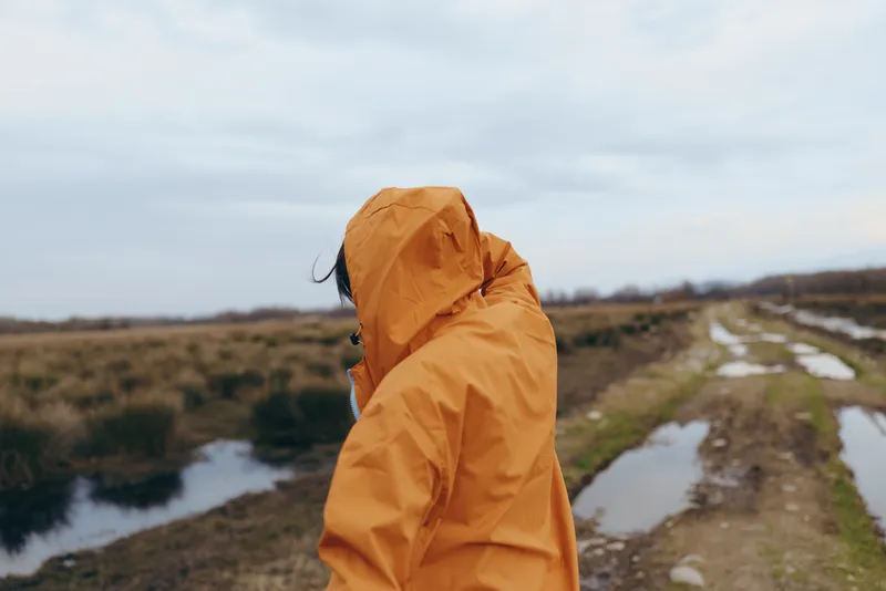 Woman lifestyle outdoors in rain jacket, hood up, walking on hiking trail by river, nature landscape with wet fields, windy sky, soft daylight, peaceful focus