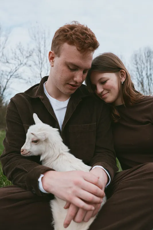 A young couple sits closely outdoors holding a white baby goat while sharing a tender moment in casual autumn clothing on a cloudy day in a natural setting.