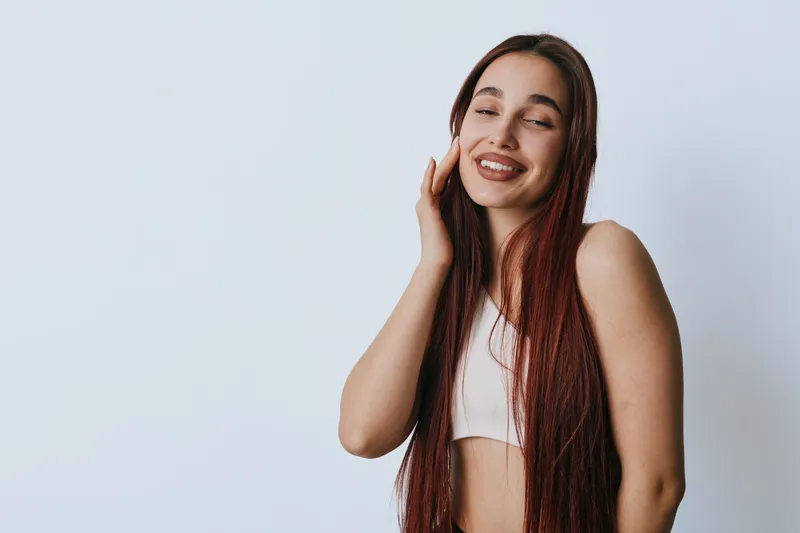Young woman with long hair smiling with closed eyes and hand touching face, wearing white top with natural makeup on light background for beauty and happiness concept.