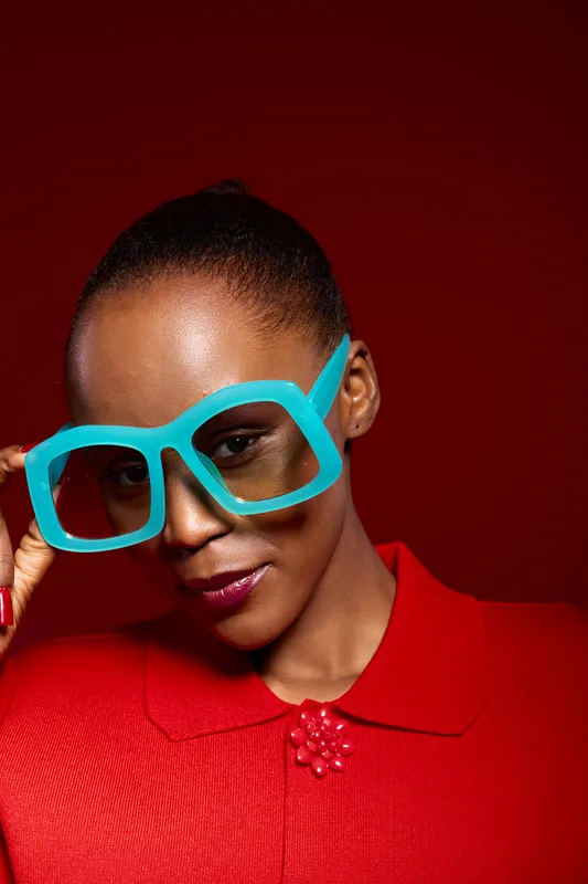 Portrait of a confident African woman with dark skin wearing large blue glasses and a red textured top against a red background. Concept of fashion, style, and beauty.
