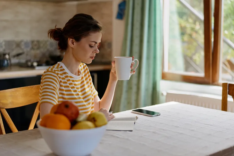 Calm woman in striped shirt sits at table with fruit bowl, mug in hand, morning light warming a cozy kitchen. A simple, bright scene emphasizing home lifestyle and everyday wellness.