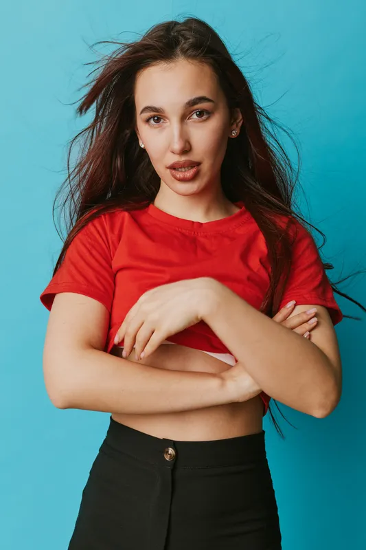 Young woman with long hair wearing red shirt and black pants posing with crossed arms against blue background. Casual style portrait with confident expression and natural makeup.