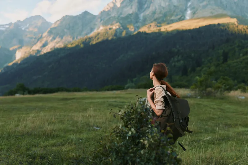 A woman with a backpack stands in a grassy meadow, looking toward distant mountains, embodying outdoor life, hiking, and solitude in nature.