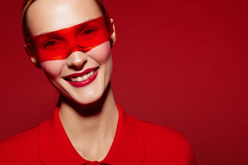 Smiling woman with red sunglasses and red shirt poses against red background. Bright makeup and glossy lips create bold fashion look. Concept of style, confidence, and modern beauty.