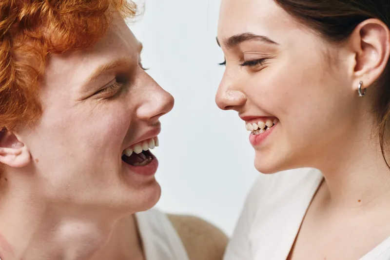 Young man and woman laughing together facing each other with happy expressions and close intimacy in studio environment. Friendship, connection, joy and positive emotion between people.