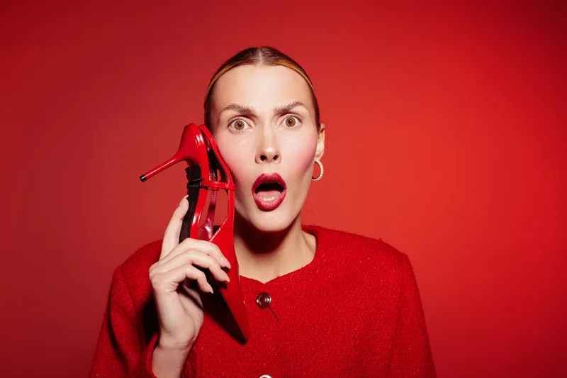 Surprised woman in red sweater holds red high heel shoe near face on red background. Concept of fashion, shock, and creative expression with vibrant color contrast.