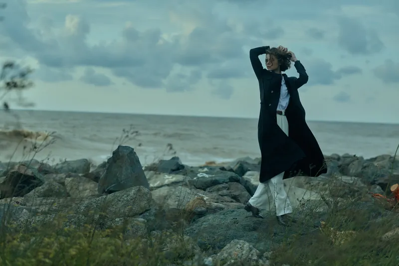 Windy coastal scene with a woman in a long coat standing on rugged rocks by the sea, captured in a dynamic pose as clouds roll over the horizon and spray hints at the harsh yet beautiful weather