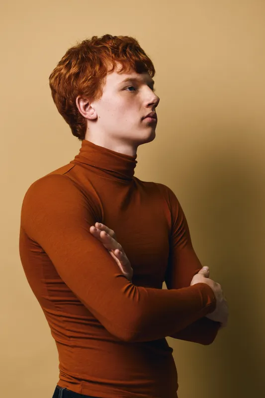 Portrait of young man with red hair wearing brown turtleneck, standing with arms crossed and looking sideways against plain beige background indoors.
